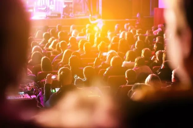 An audience seen from behind, lit by warm stage lights.