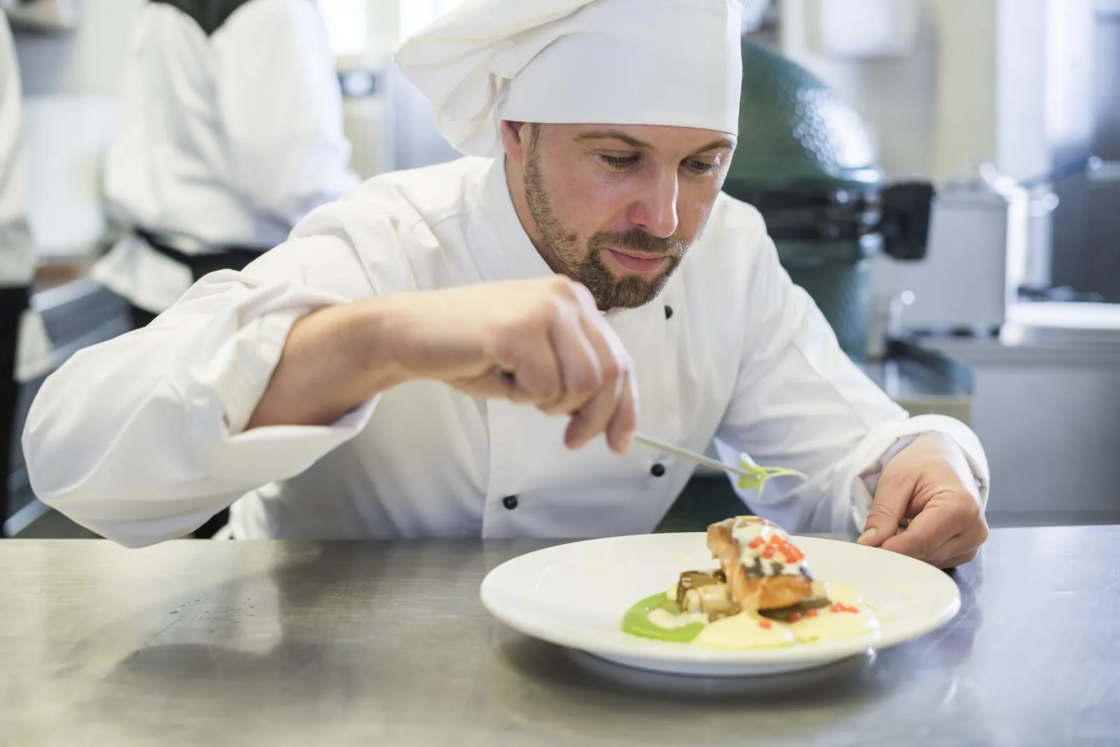 A chef in whites finishes a plated dish at a kitchen pass.