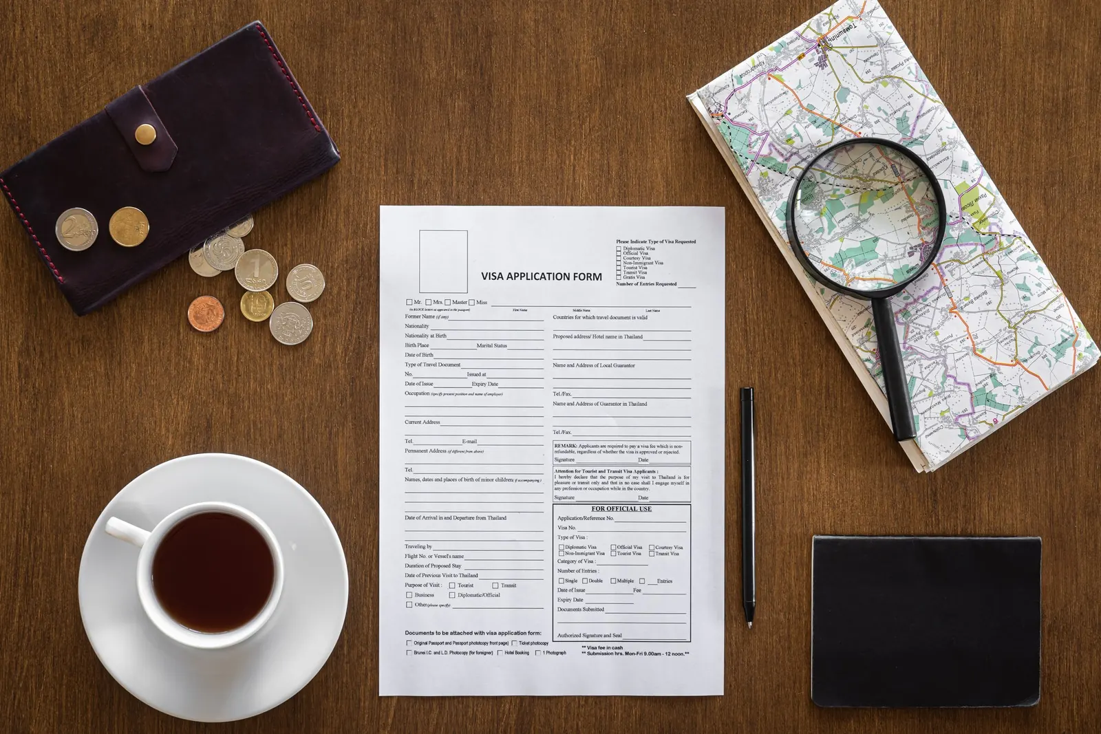 A visa application form on a wooden table beside a passport, a map and coins.